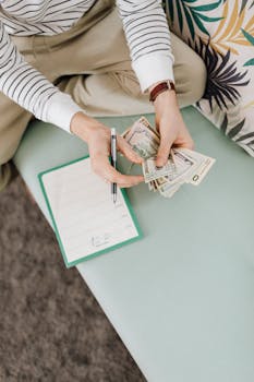 An individual counts money while sitting, holding a notebook and pen.