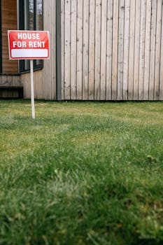 Red 'House for Rent' sign on green lawn with wooden house wall in background.
